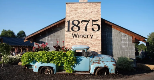 The rustic exterior of 1875 Winery showcases a vintage blue pickup truck filled with greenery parked in front of a brick chimney featuring the winery's logo.