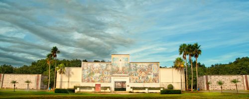 A large mural on a building surrounded by palm trees, set against a cloudy sky. Green grass in the foreground.
