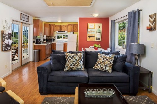 Cozy living room with a dark sofa, decorative pillows, and an open kitchen area featuring wooden cabinets and a dining table.