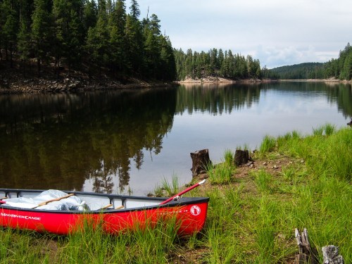 A red canoe rests on the shore of a calm lake, surrounded by green grass and trees under a cloudy sky.
