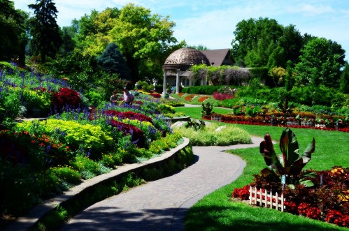 A vibrant garden with colorful flowers, winding paths, and a gazebo in the background under a clear blue sky.