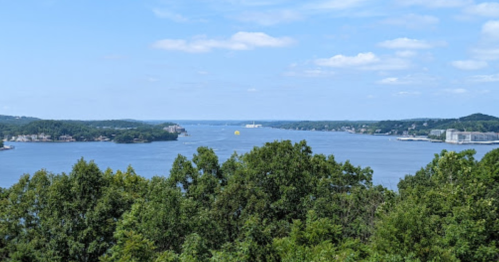 A scenic view of a lake surrounded by green trees and distant hills under a clear blue sky.