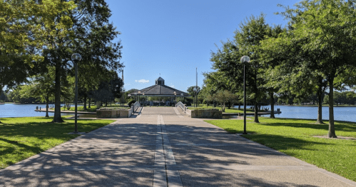 A paved path leads to a gazebo by a lake, surrounded by trees and green grass under a clear blue sky.