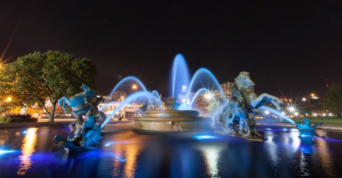 A nighttime view of a fountain with illuminated water jets and sculptures of horses and figures, surrounded by trees.