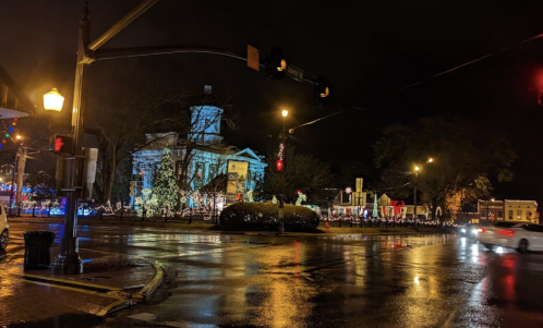 A nighttime street scene with a historic building illuminated, wet pavement reflecting lights, and traffic signals visible.