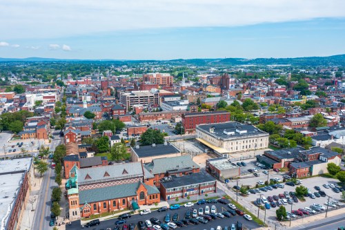 Aerial view of a vibrant cityscape with buildings, greenery, and parked cars under a clear blue sky.