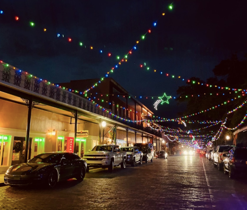 A vibrant street at night adorned with colorful lights and festive decorations, lined with parked cars.