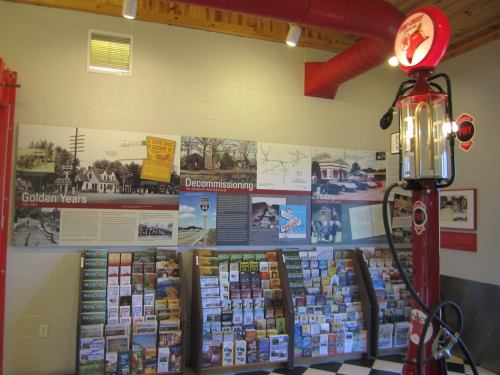 A vintage gas pump stands beside a display of brochures and historical photos on a wall in a museum-like setting.
