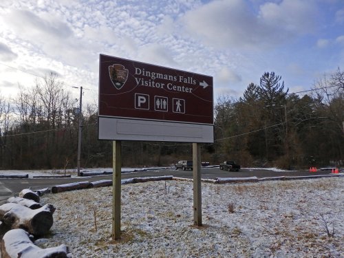 Sign for Dingmans Falls Visitor Center with parking and hiking symbols, surrounded by snow and trees.