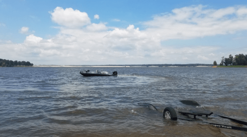 A boat on a calm lake under a partly cloudy sky, with a trailer partially submerged in the foreground.