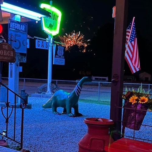 A green dinosaur statue in front of a gas station, with fireworks in the night sky and an American flag nearby.