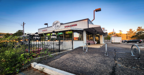 A modern café with outdoor seating, surrounded by greenery, under a clear blue sky.