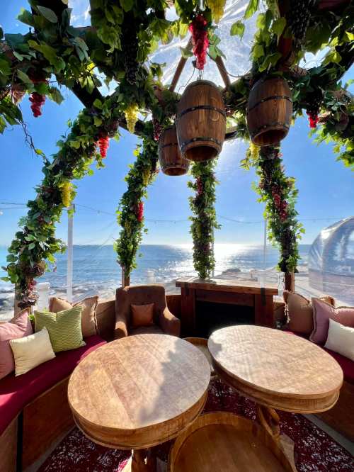 Cozy seating area with wooden tables, surrounded by greenery and barrels, overlooking a scenic ocean view.