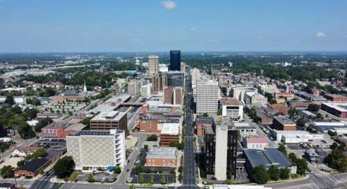 Aerial view of a city skyline featuring tall buildings, roads, and green spaces under a clear blue sky.