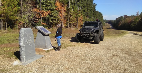 A person stands by a historical marker near a dirt road, with a black off-road vehicle parked nearby and trees in the background.