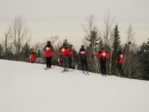 A group of skiers in red jackets with white crosses stands on a snowy slope, surrounded by trees.