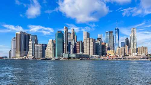 A panoramic view of a modern city skyline with tall buildings against a blue sky and water in the foreground.