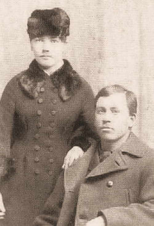A vintage black-and-white photo of a man seated and a woman standing beside him, both dressed in formal attire.