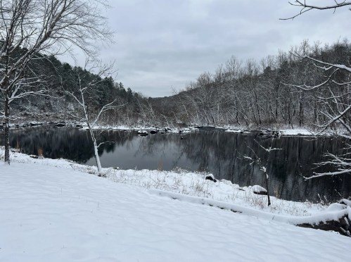 A serene winter landscape featuring a snow-covered riverbank and trees, with a calm, reflective water surface.