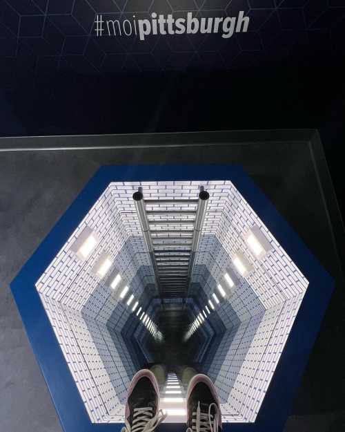 A person’s feet stand on a glass floor overlooking a deep, illuminated hexagonal shaft with a tiled interior.