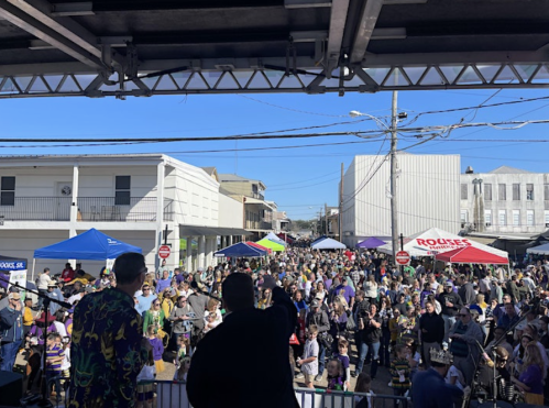 A lively crowd gathers at a festival, with colorful tents and a clear blue sky in the background.