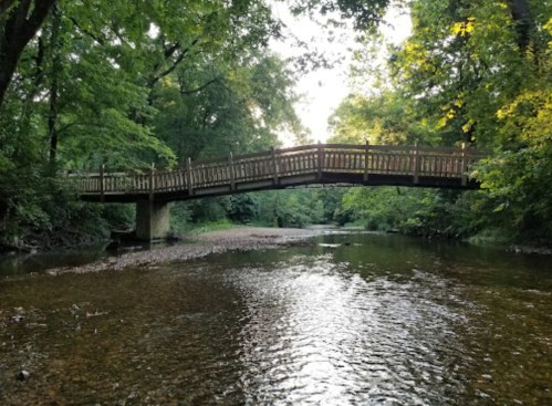 A wooden bridge spans a calm river, surrounded by lush green trees and dappled sunlight.