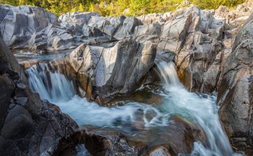 A serene river flows over smooth, rocky terrain, surrounded by lush greenery and sunlight filtering through the trees.