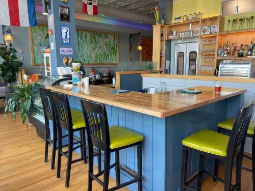 A cozy café interior featuring a wooden bar with green stools, plants, and colorful decor in a bright atmosphere.