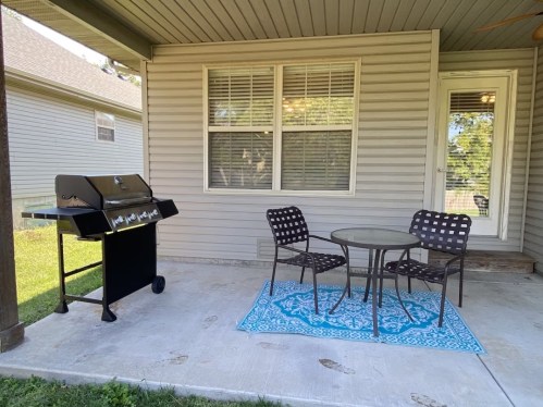 A patio with a grill, a round table, and two chairs on a blue rug, surrounded by a house and greenery.