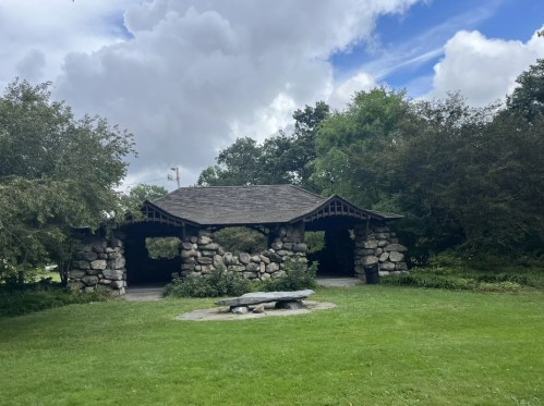 A stone pavilion with a wooden roof surrounded by greenery and a grassy area under a partly cloudy sky.