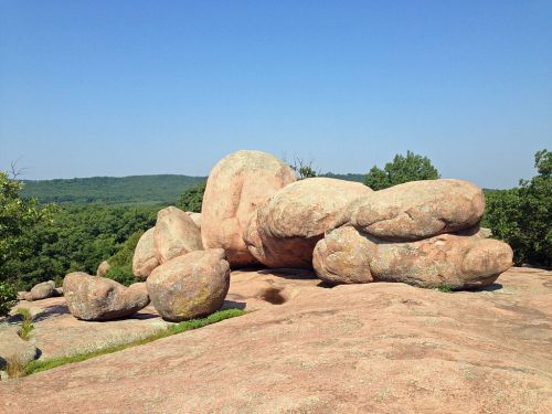Large, smooth boulders resting on a rocky surface, surrounded by green trees and a clear blue sky.
