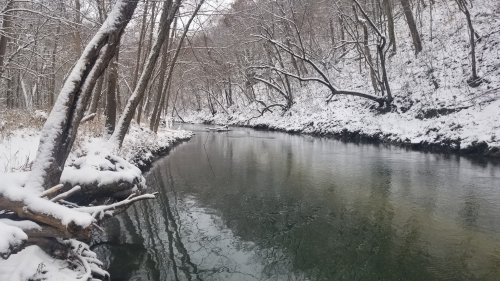 A serene winter scene of a snow-covered riverbank with trees and calm water reflecting the snowy landscape.