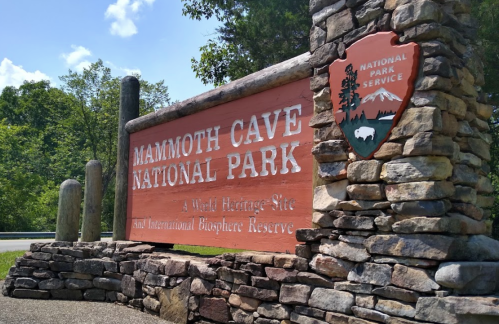 Sign for Mammoth Cave National Park, featuring stone structure and park emblem, surrounded by greenery and blue sky.