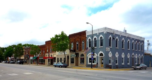 A street view of a small town featuring brick buildings, shops, and trees under a cloudy sky.