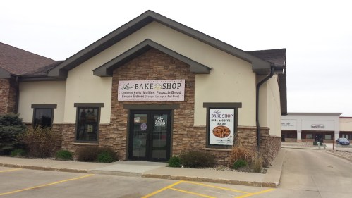 Exterior of a bakery shop with a stone facade, featuring a sign that reads "Bake Shop" and advertisements for baked goods.