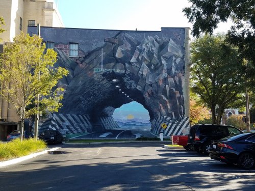 A large mural depicting a tunnel with a sunset and mountains, surrounded by trees and parked cars.