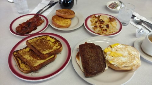A breakfast spread featuring French toast, bacon, hash browns, and a sandwich with egg on a bagel.
