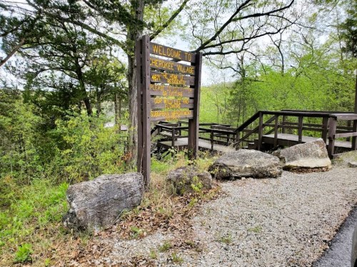 A wooden sign welcomes visitors to a village, surrounded by greenery and rocky terrain. Pathways are visible nearby.