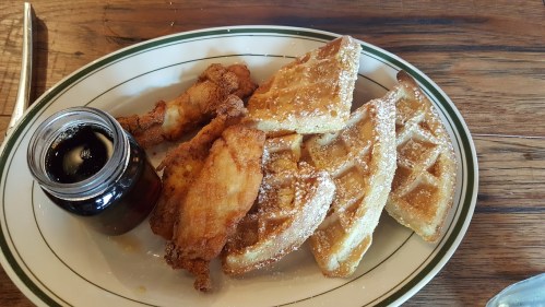 A plate of crispy fried chicken served with golden waffles and a small jar of syrup.