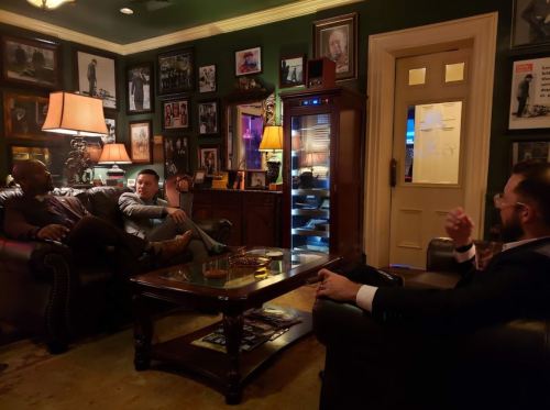 Three men in a cozy, dimly lit room with vintage decor, engaged in conversation near a coffee table and a glass display.