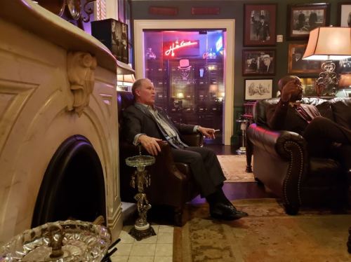 A man sits in a vintage armchair by a fireplace, while another man relaxes on a leather couch in a cozy, dimly lit room.