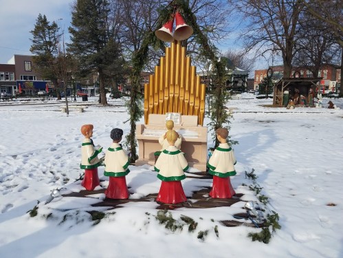 A winter scene featuring a decorative organ with carolers in festive attire, surrounded by snow and trees.