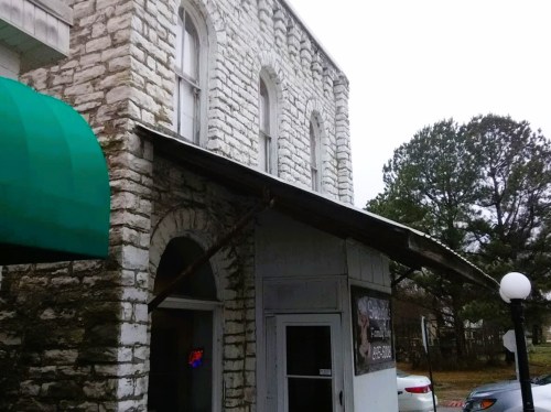 Historic stone building with a green awning and a sign, surrounded by trees on a cloudy day.
