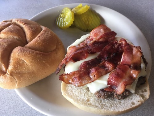 A plate with a burger topped with bacon and mayonnaise, alongside a bun and pickles.