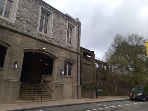 A stone building with stairs, under a cloudy sky, next to a road with a parked vehicle and trees in the background.