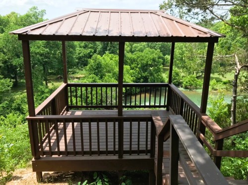 A wooden gazebo with a metal roof overlooks a lush green landscape and a winding river below.
