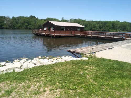 A wooden dock extends over a calm lake, with a small cabin on the water surrounded by greenery and clear blue skies.
