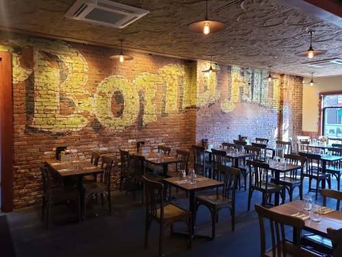 Interior of a restaurant featuring exposed brick walls with "BOMBAY" painted in large letters and wooden tables.