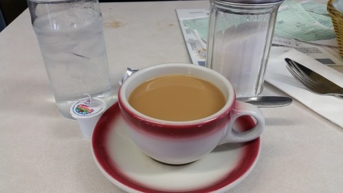 A cup of coffee with cream sits on a table next to a glass of water, a sugar container, and a fork.