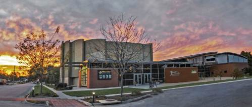 A modern building at sunset, with vibrant clouds and trees in the foreground, showcasing a welcoming entrance.
